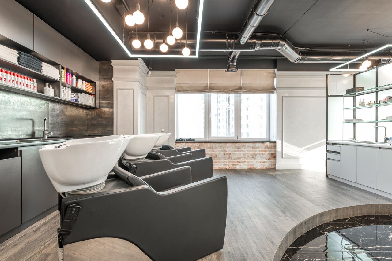 Black backwash chairs with sinks placed in modern spacious beauty salon with window and arrangement of cosmetology supply on shelves